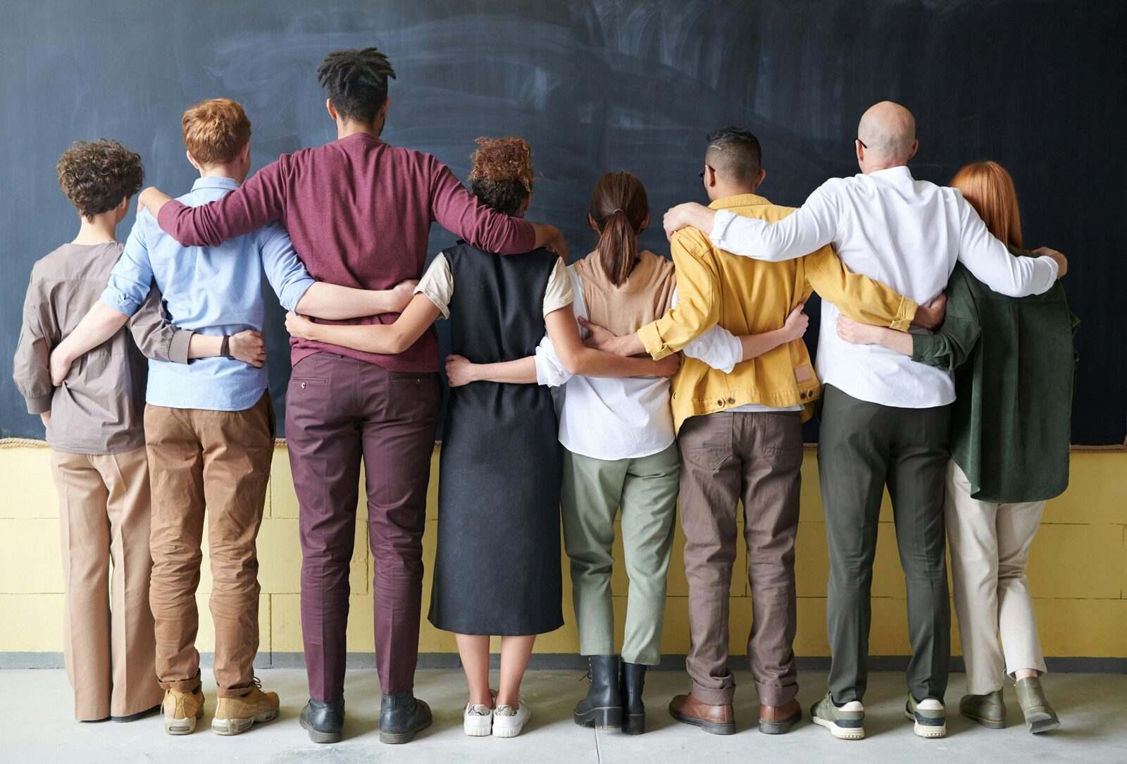 A diverse group of students stand with their backs facing the camera, looking at a chalkboard, arms around one another's shoulders.