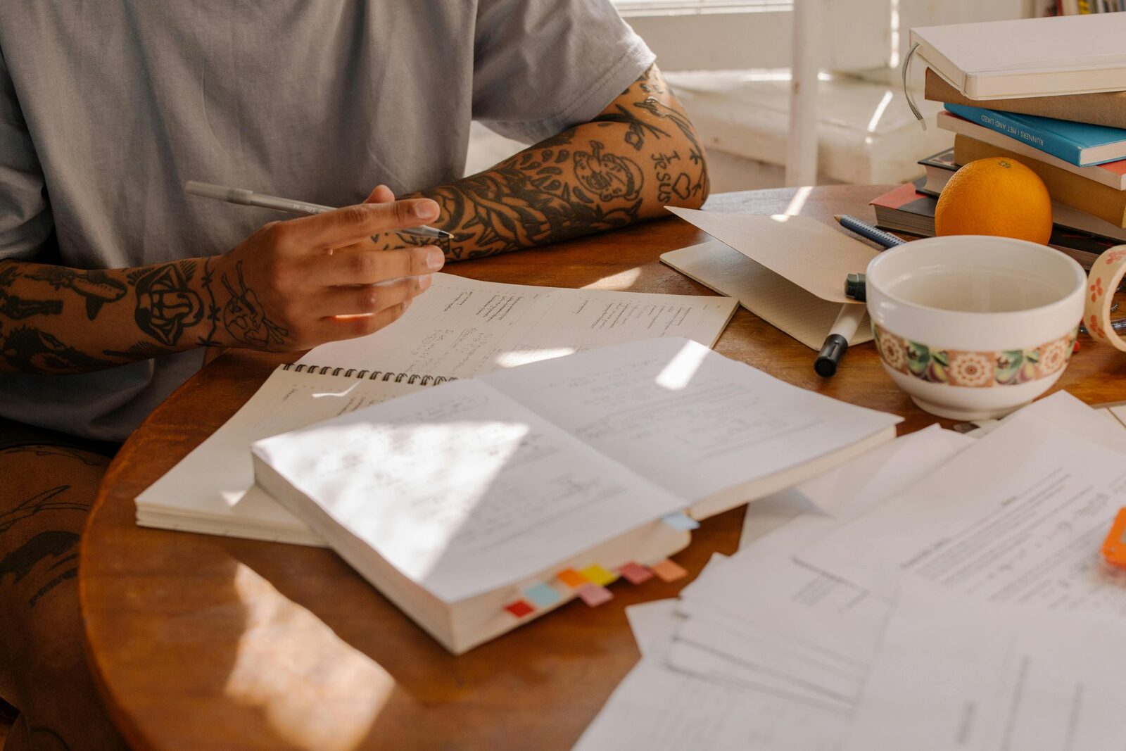 A man with tattooed arms sits at a table with pen in hand and several notebooks open in front of him.