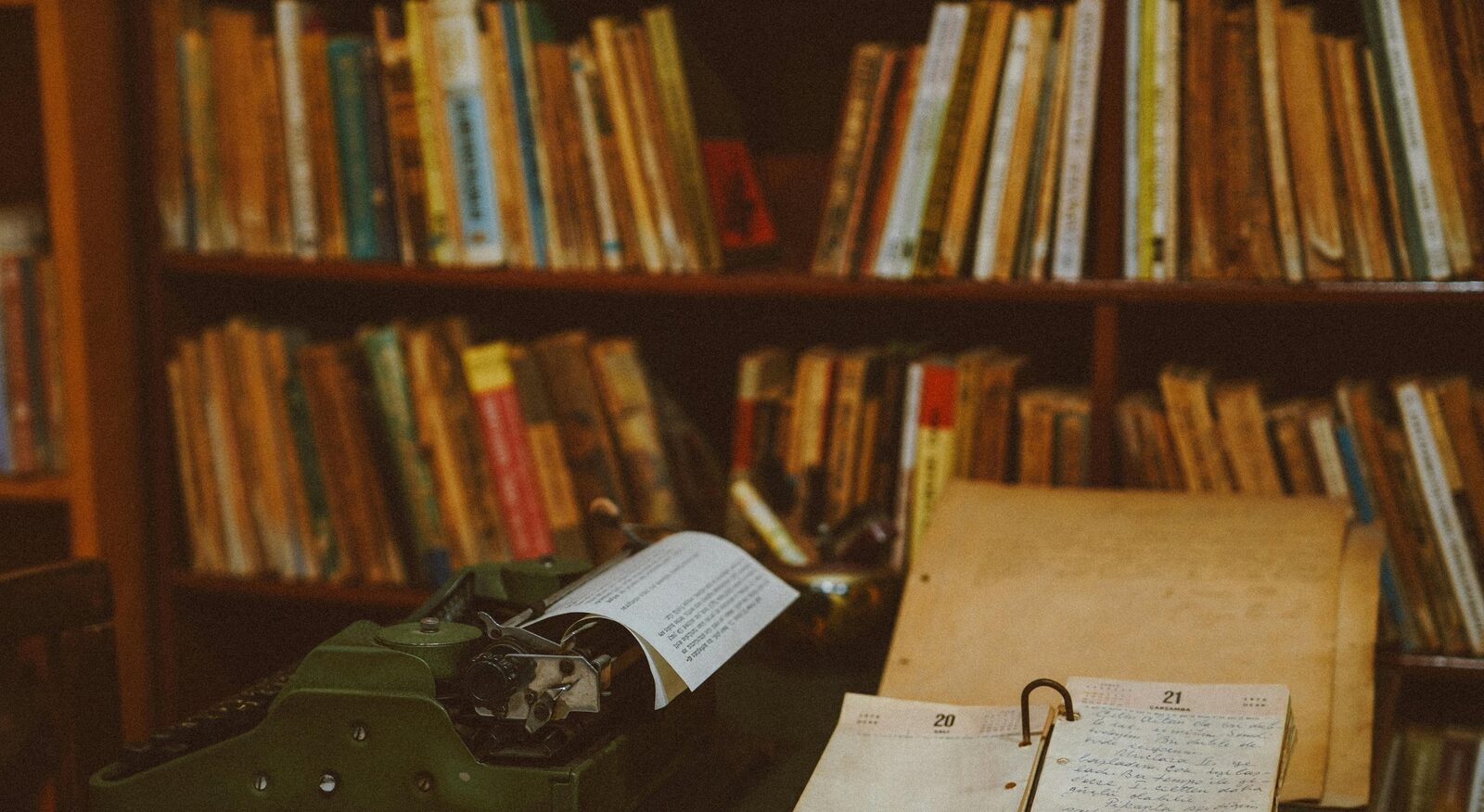 A writing desk with a typewriter, handwritten pages, and a paper calendar, set against a full bookcase.